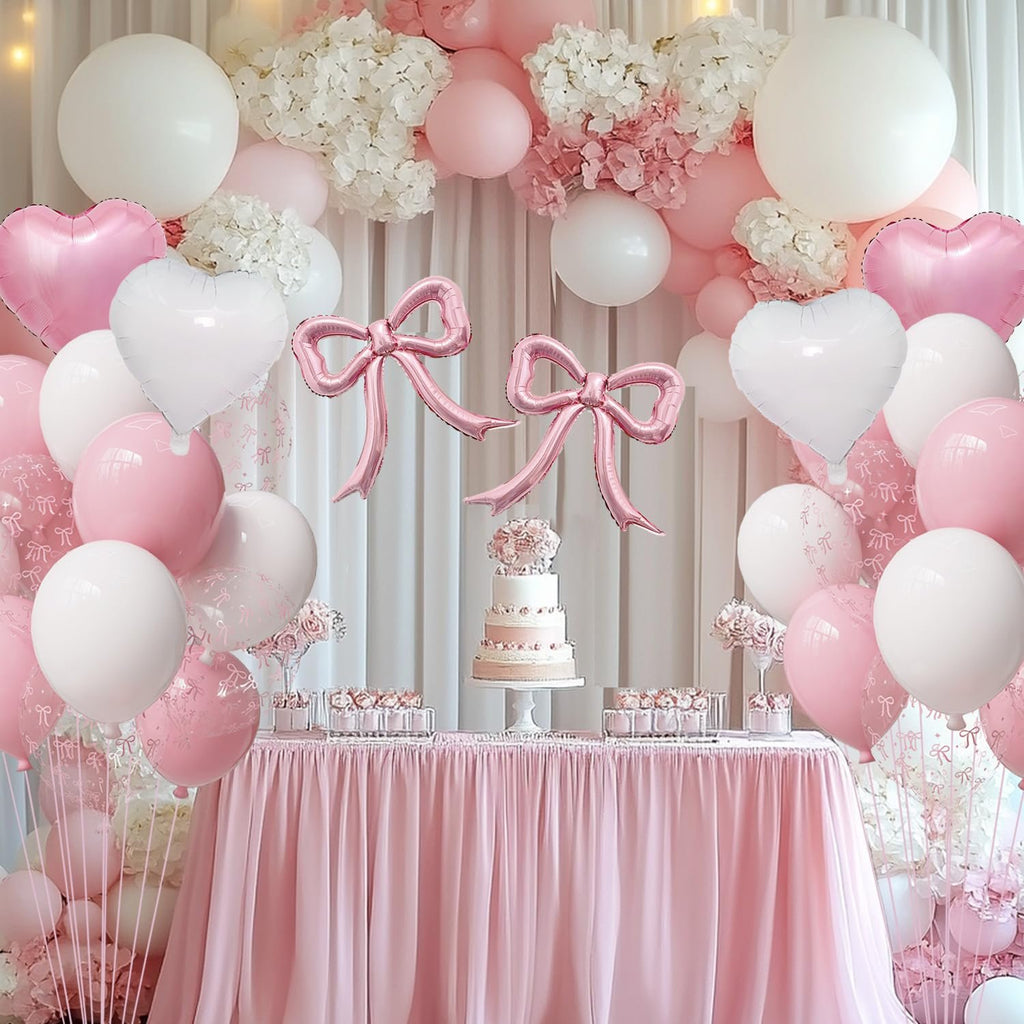 Pink and white balloon arrangement with bow-shaped and heart-shaped foil balloons, set against a draped backdrop with a cake on a table.