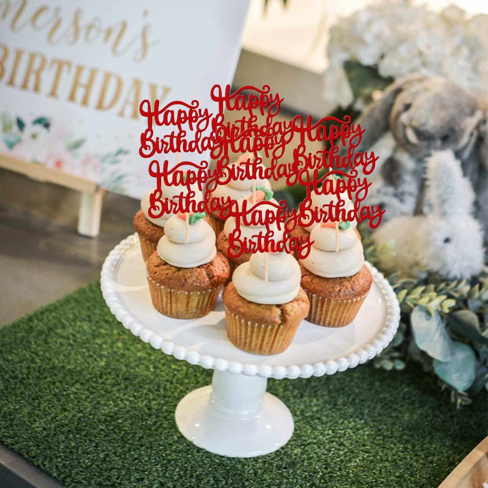 Red glitter 'Happy Birthday' cupcake toppers inserted into frosted cupcakes on a white cake stand, with a blurred background.