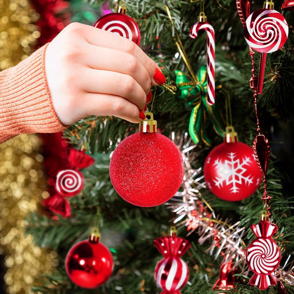 Hand decorating a Christmas tree with red and white shatterproof ornaments in ball, candy, and lollipop shapes.