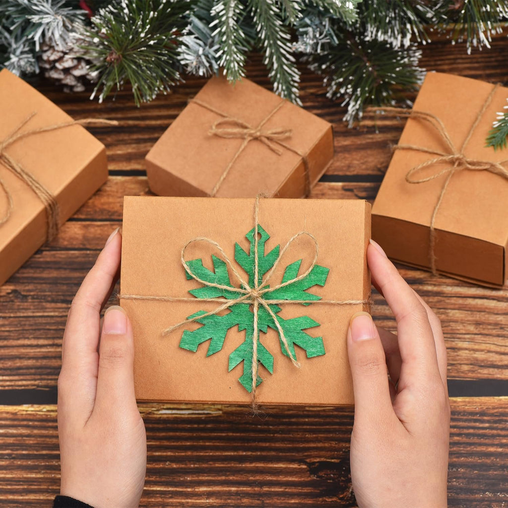 Hands holding a brown gift box decorated with a green snowflake ornament and jute twine, surrounded by Christmas greenery and other gift boxes.