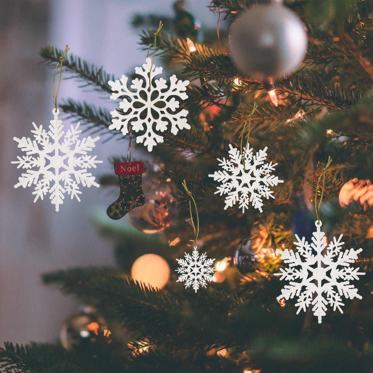 White glitter snowflake ornaments of various sizes hang on a Christmas tree with bokeh lights.