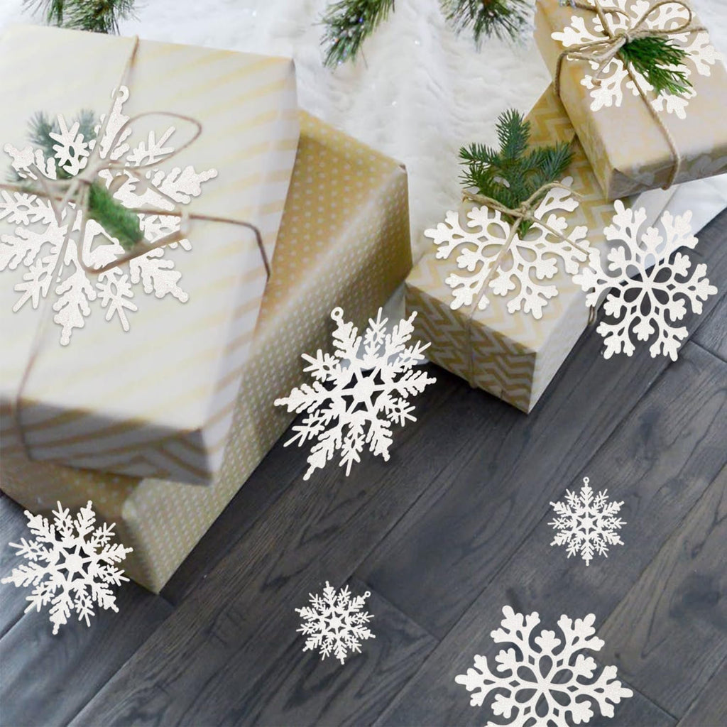 White glitter snowflake ornaments scattered around gift-wrapped presents on a wooden floor with pine branches.