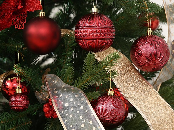 Assortment of wine red shatterproof Christmas ball ornaments in various sizes and embossed patterns hanging on a decorated evergreen tree with gold and patterned ribbon.