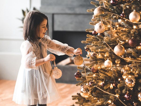 Young girl in a white dress decorating a Christmas tree with wine red and gold ornaments.