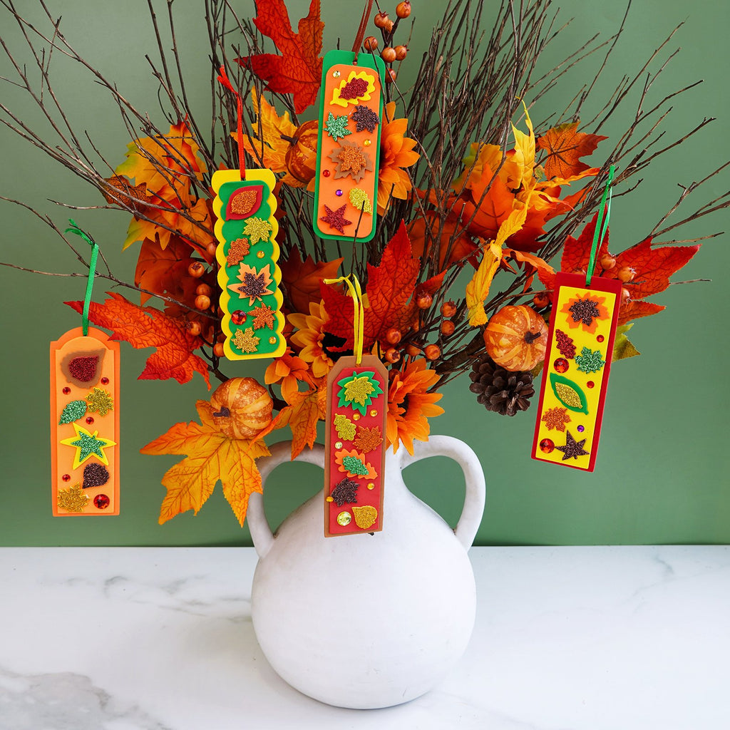 Five DIY fall leaf bookmarks with glitter and gems displayed in a vase with autumn foliage