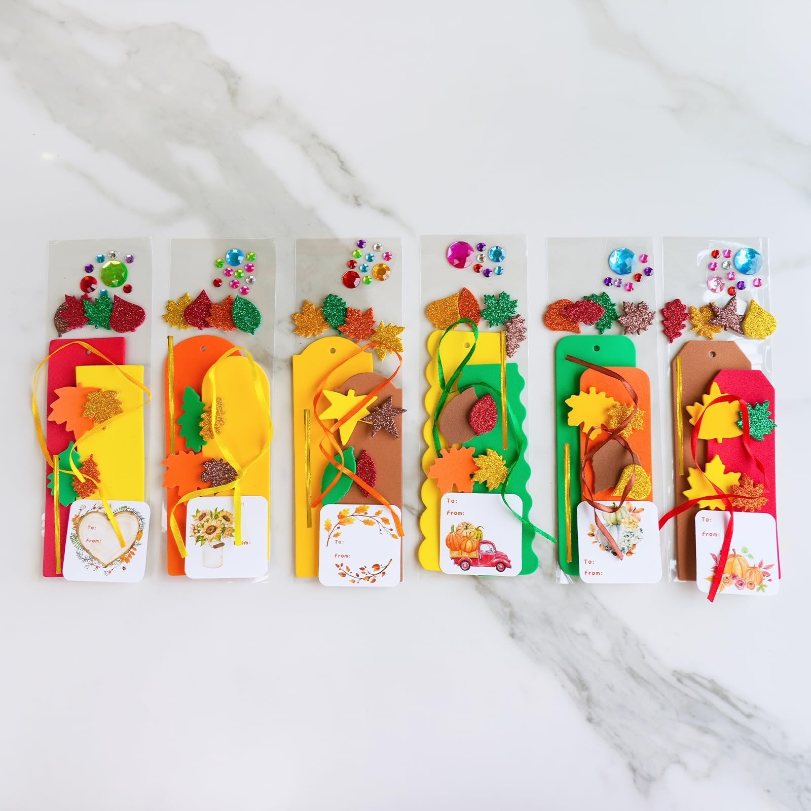 Six fall craft kits for making leaf bookmarks, each with foam shapes, glitter stickers, and ribbons on a marble background.