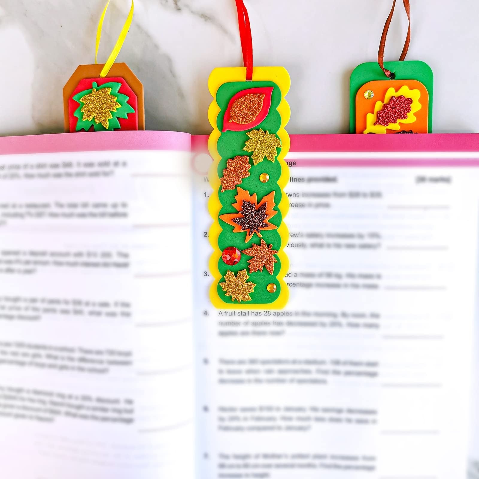 Three DIY fall leaf bookmarks made of foam and glitter, placed in an open book.