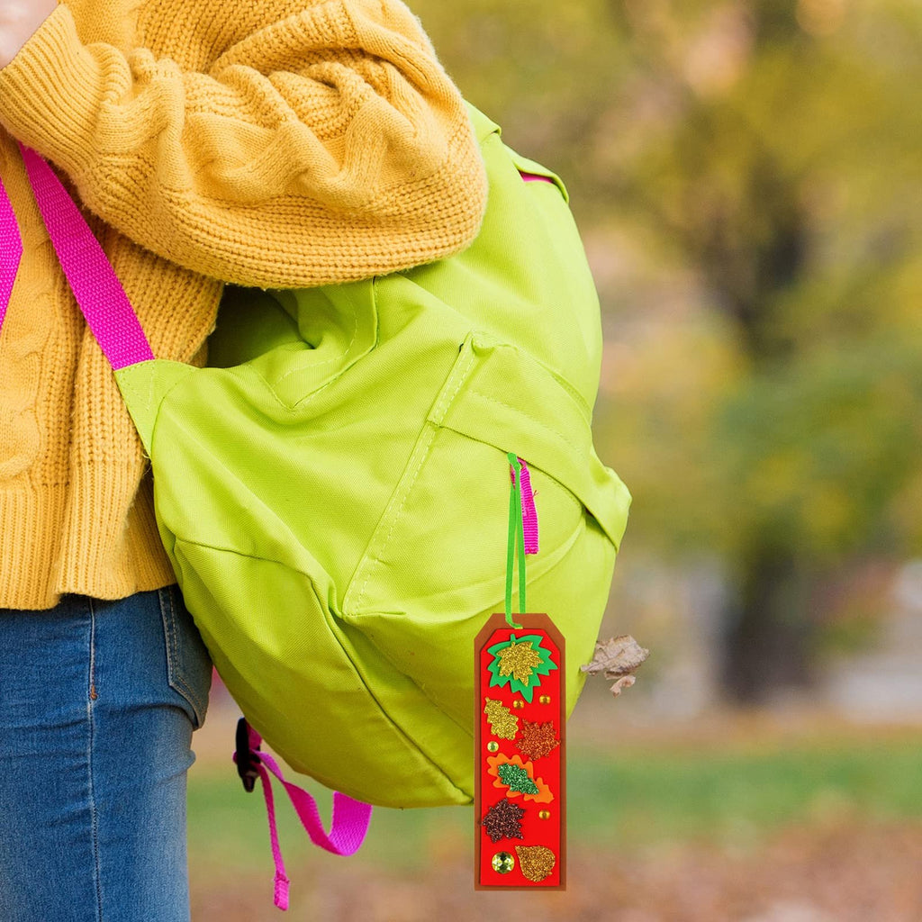 Person wearing yellow sweater and jeans, carrying a green backpack, with a handmade fall leaf bookmark hanging from it.