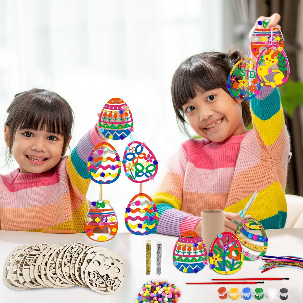Two smiling girls hold up colorful Easter egg ornaments they have decorated, with craft supplies laid out on the table.
