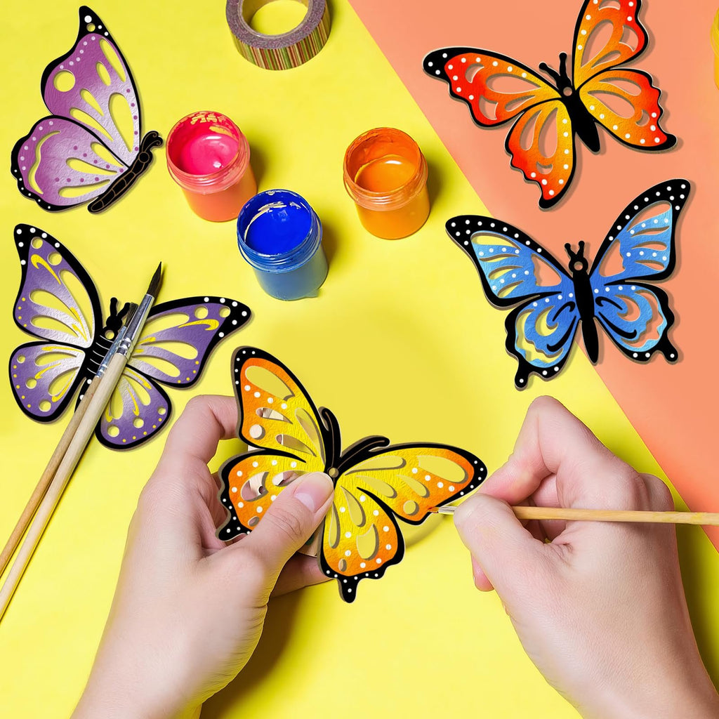 Hands painting unfinished wooden butterfly ornaments with paint pots and brushes on a yellow and orange background.