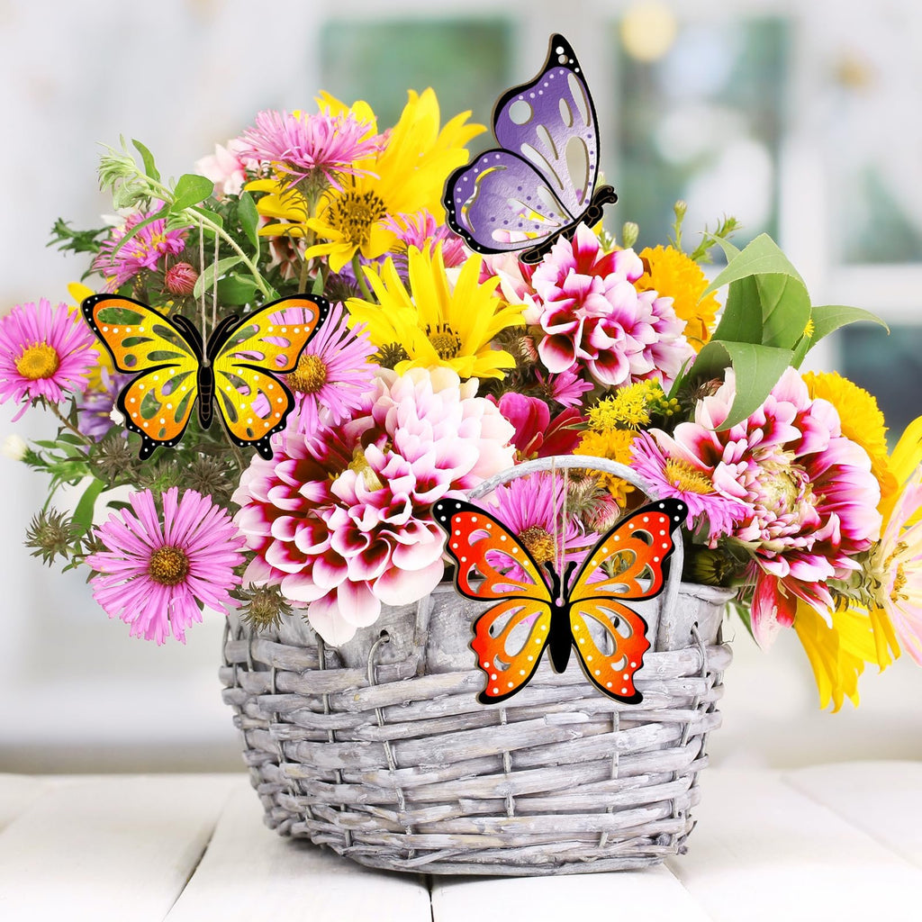 Three unfinished wooden butterfly ornaments hanging in a basket of colorful flowers on a white wooden surface.