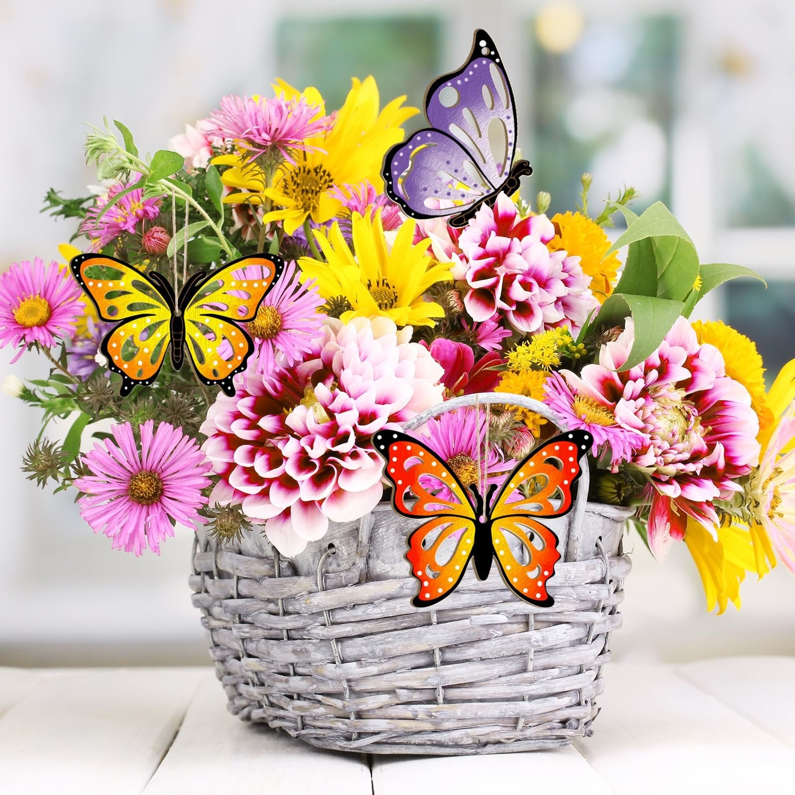 Three unfinished wooden butterfly ornaments hanging in a basket of colorful flowers on a white wooden surface.