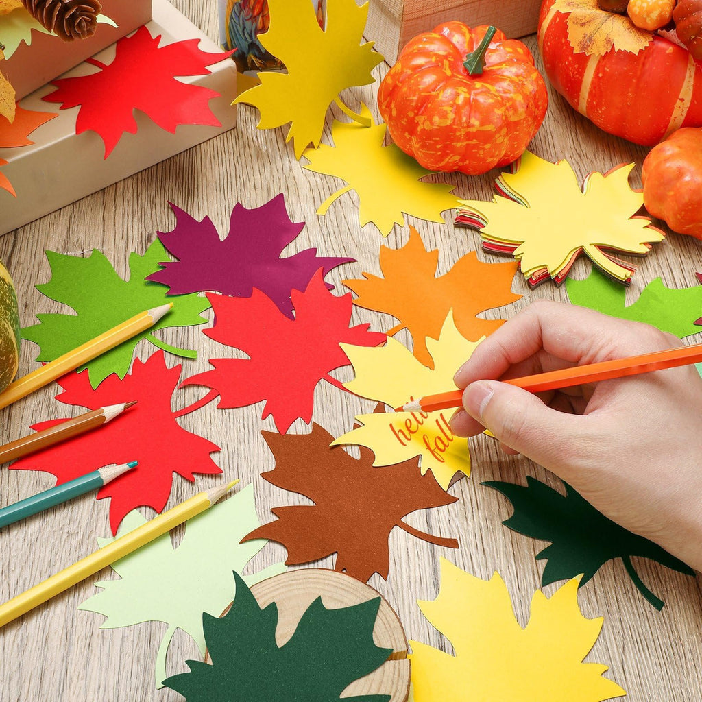 Hand coloring paper maple leaves with colored pencils, surrounded by autumn leaf cutouts and pumpkins.