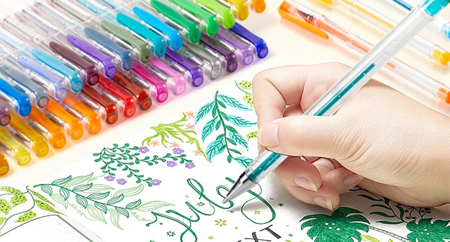 Hand holding a teal gel pen, drawing leaves and the word 'July' in a notebook, with a tray of colorful gel pens in the background.