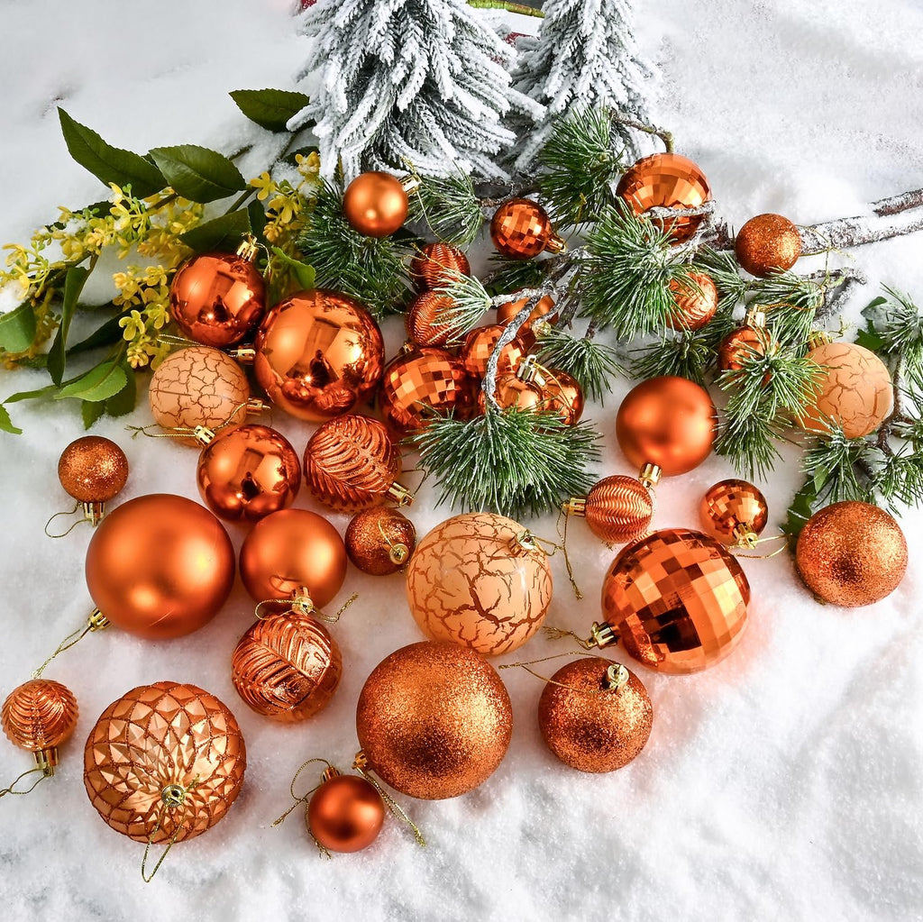 Assortment of copper orange Christmas ball ornaments in various sizes and styles scattered on artificial snow with pine branches and greenery.