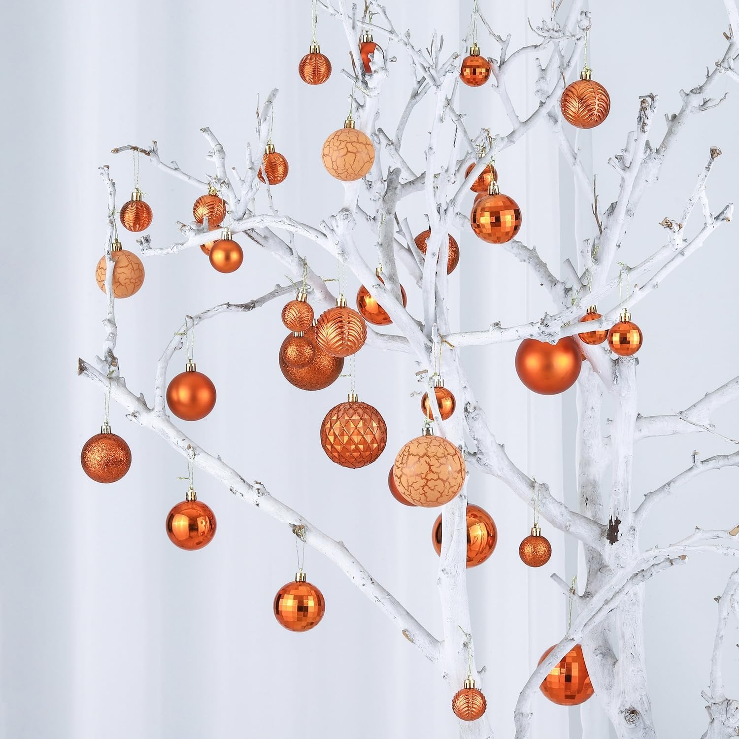 Copper orange Christmas ball ornaments in various sizes and styles hanging on a white tree branch against a white background.