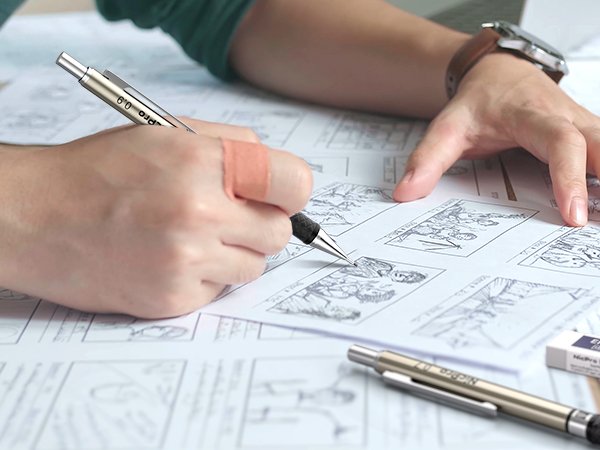 Person's hand with a band-aid drawing on storyboard paper with a mechanical pencil, another pencil and eraser nearby.