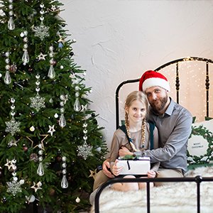 Father and daughter with Christmas tree and gift, daughter holds gift box, father wears Santa hat.