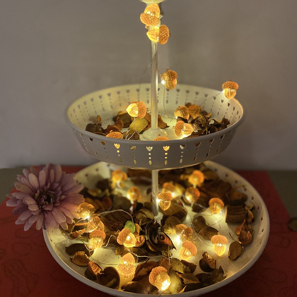 Acorn-shaped string lights with 30 LEDs displayed on a two-tiered stand filled with dried leaves and potpourri, with a pink flower on the side.