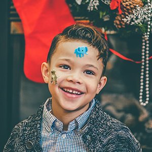 Smiling young boy with 3D puffy stickers of a dinosaur and an elephant on his face, festive background with stocking and garland