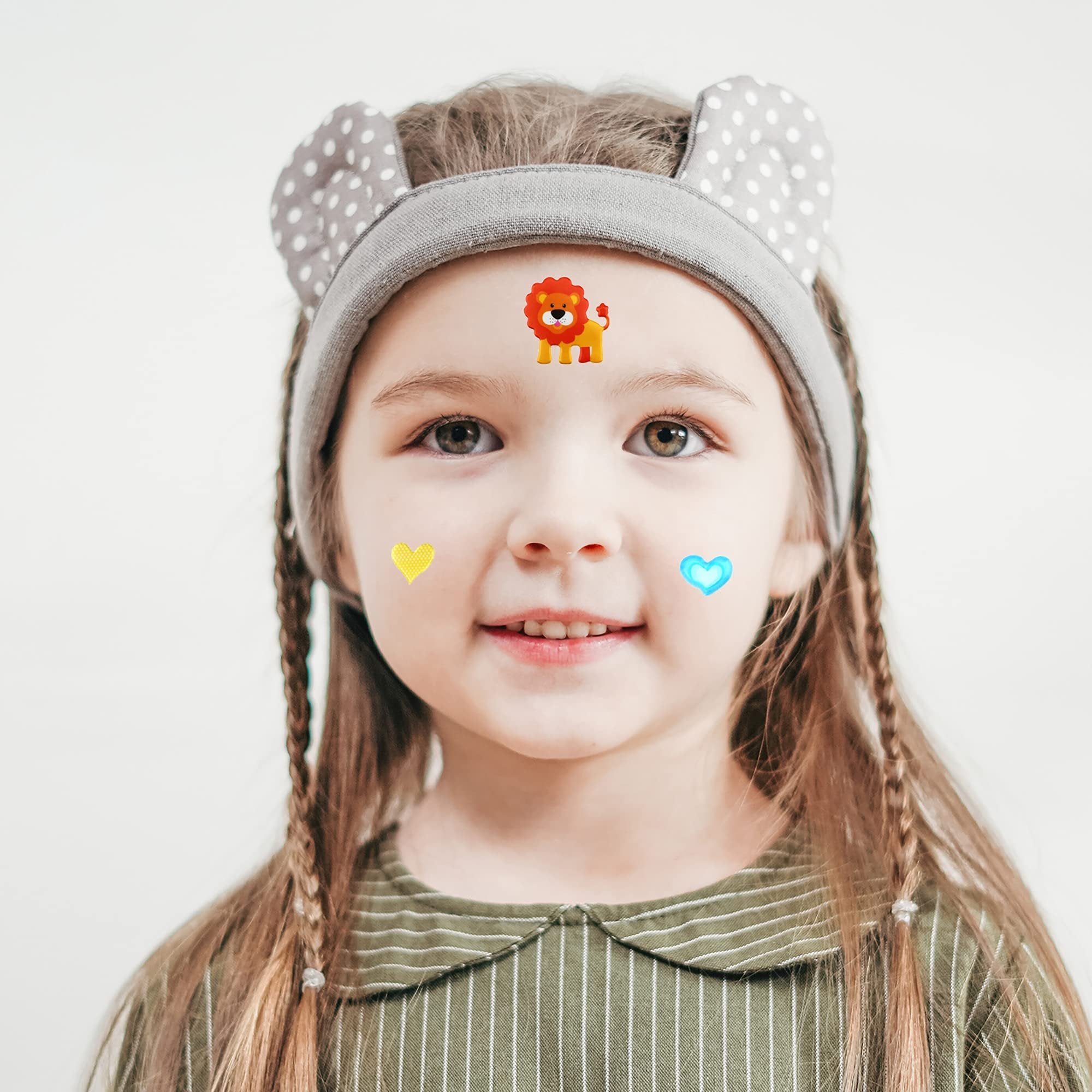 Young girl with braided hair and a headband with ears, wearing puffy stickers of a lion, a yellow heart, and a blue heart on her face.