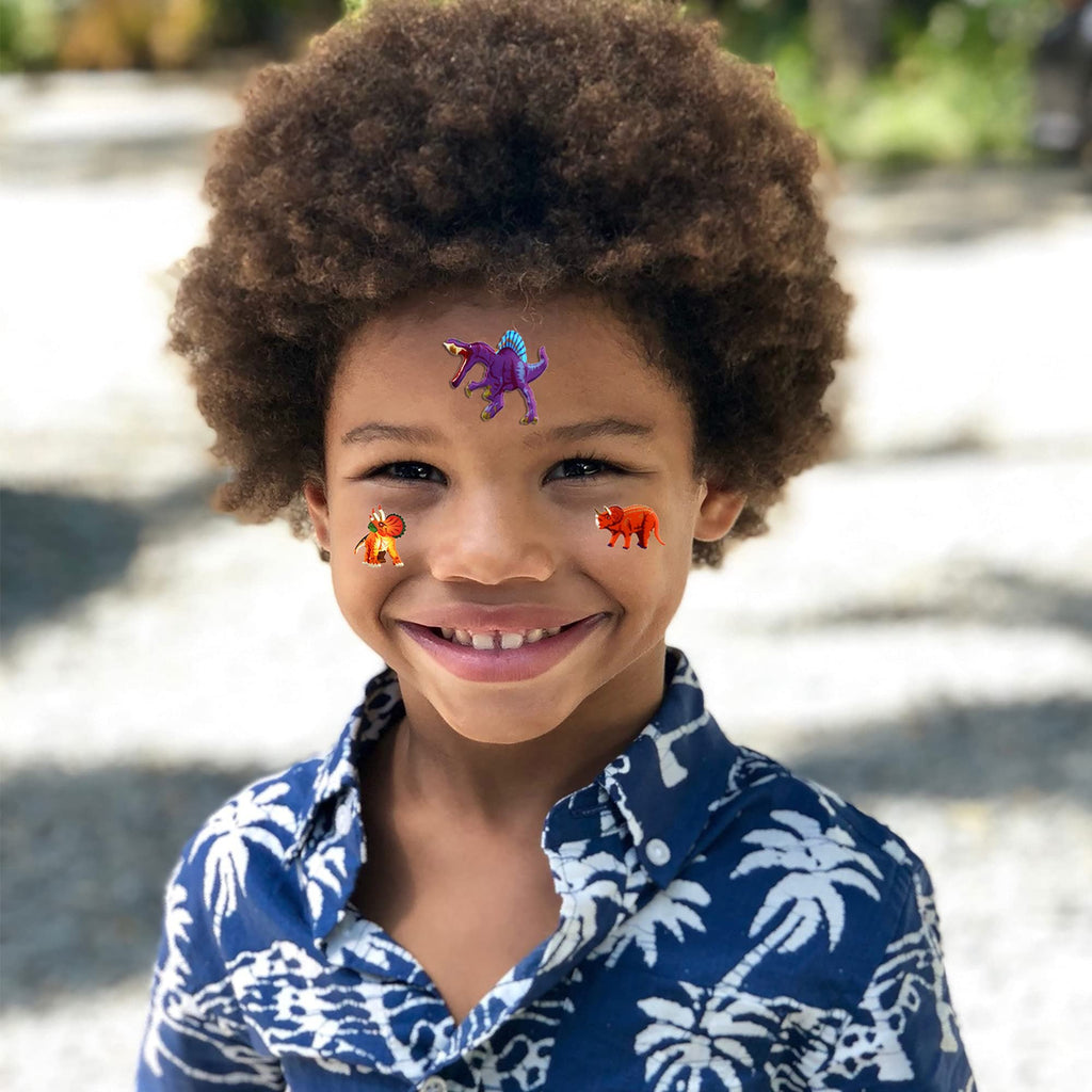 Smiling young boy with dinosaur stickers on his forehead and cheeks, wearing a blue palm tree print shirt.