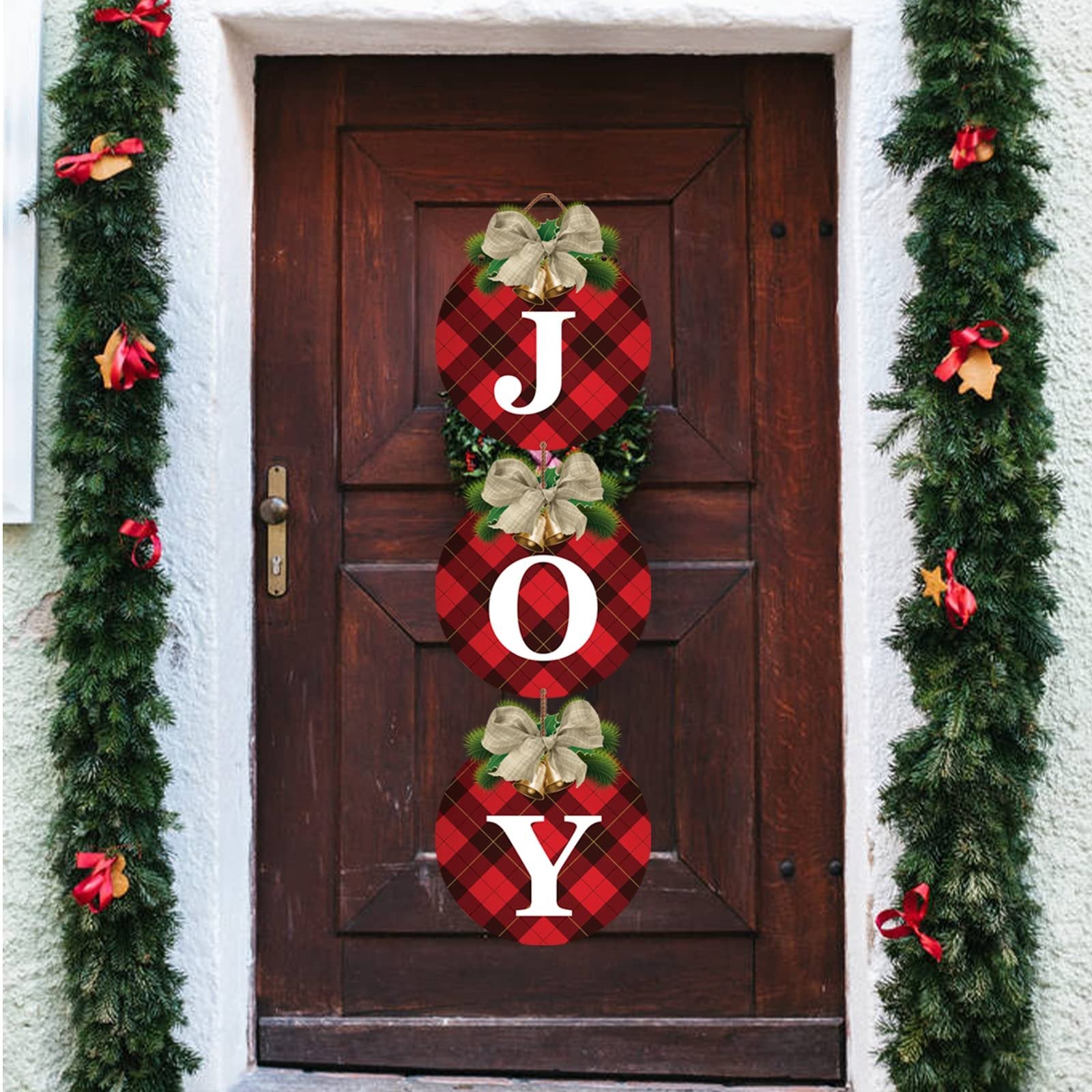 Three buffalo plaid Christmas wreath signs spelling JOY, hanging on a wooden door with evergreen garlands.