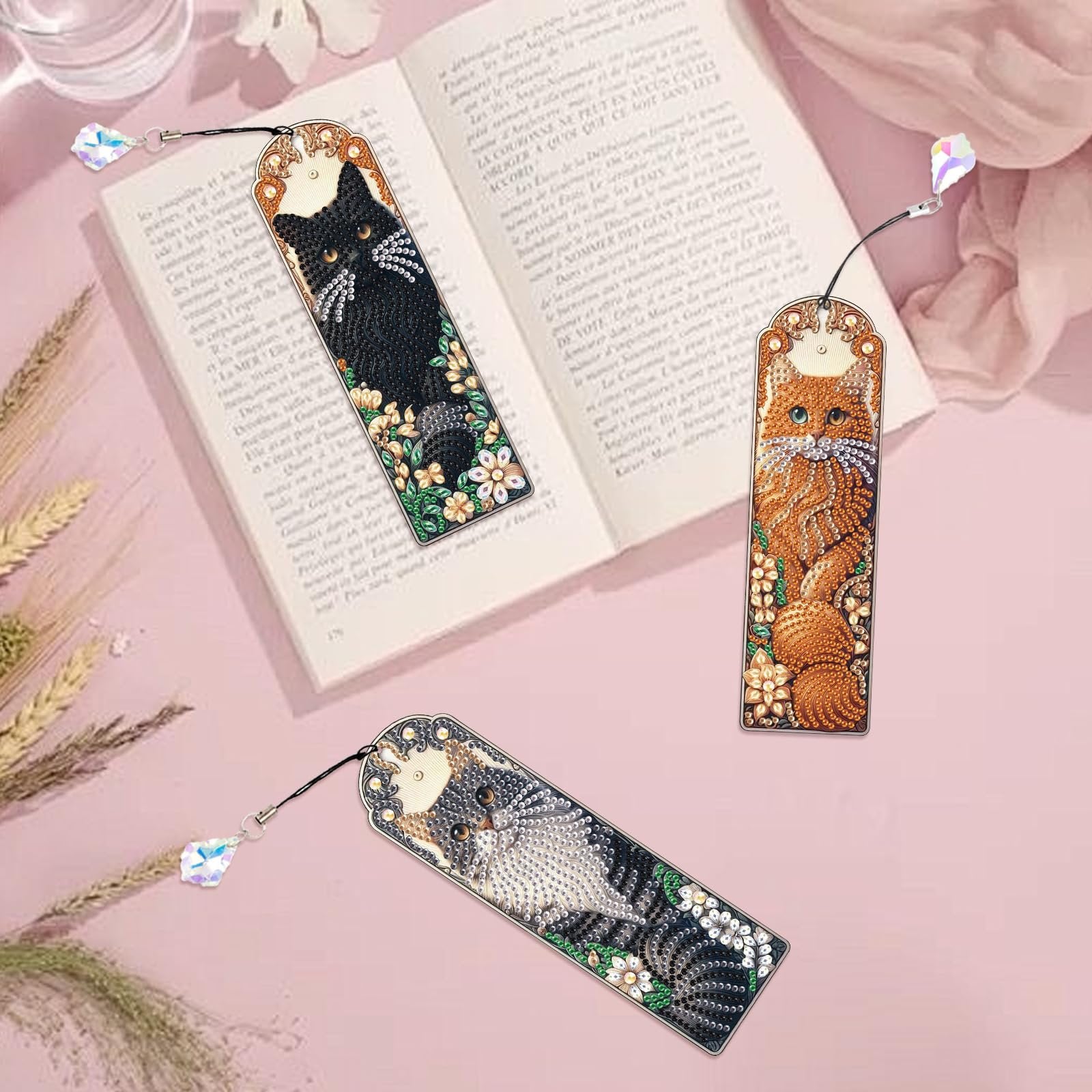 Three diamond art painting bookmarks with cat designs and crystal pendants on an open book.