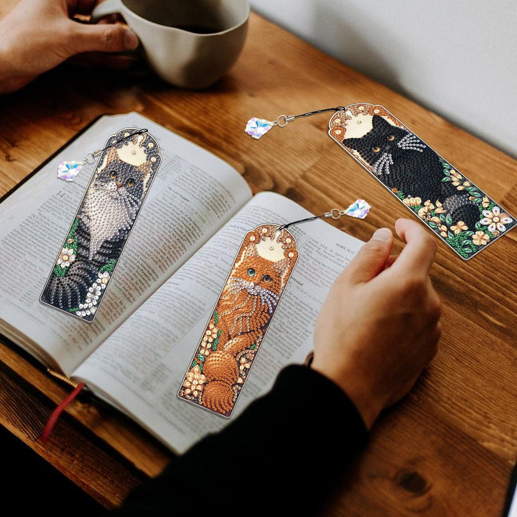 Three diamond art painting bookmarks with crystal pendants resting on an open book, featuring cat designs and floral accents.