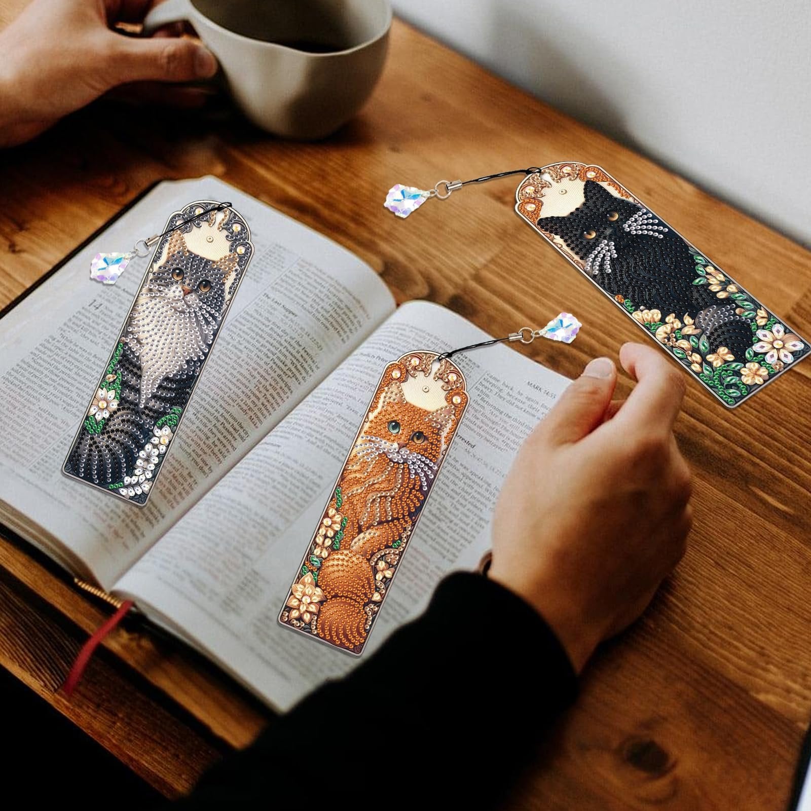 Three diamond art painting bookmarks with crystal pendants resting on an open book, featuring cat designs and floral accents.