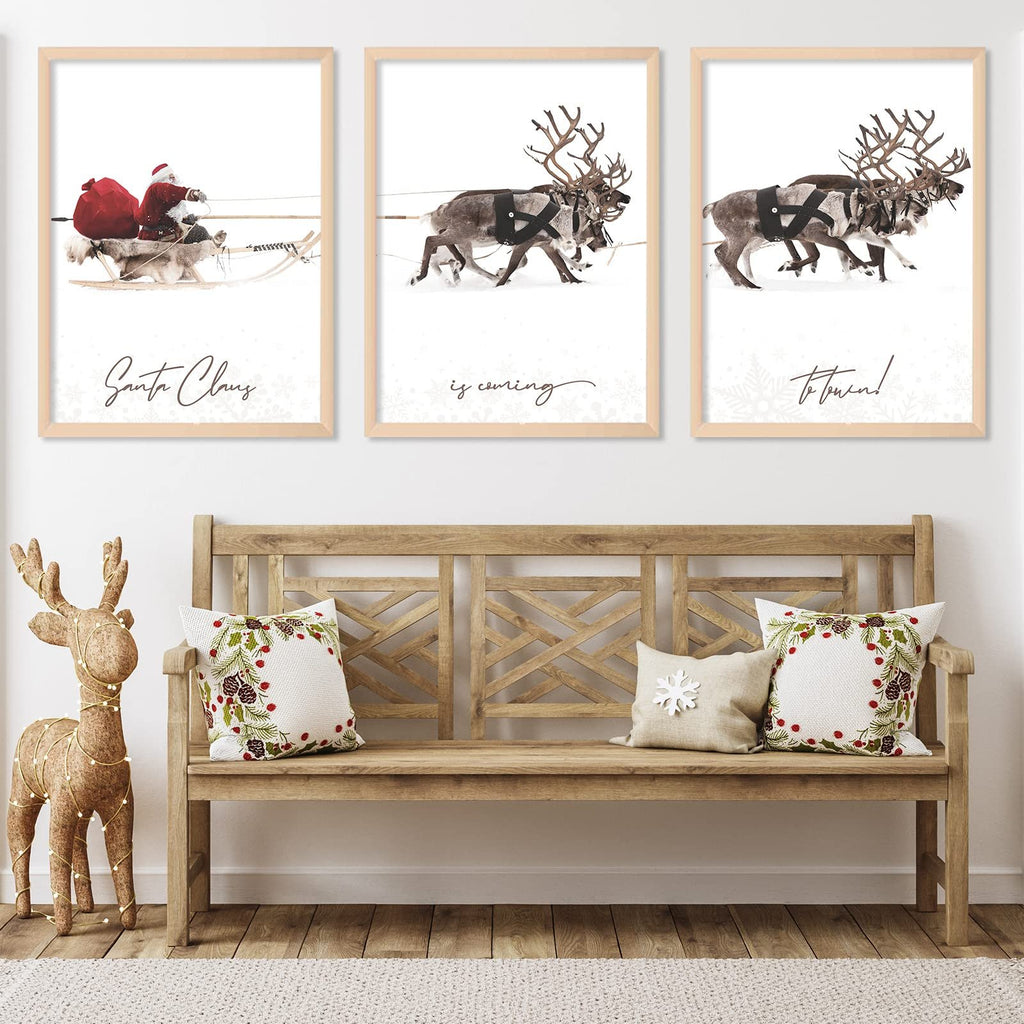 Three framed Christmas art prints depicting Santa Claus in a sleigh and reindeer pulling a sleigh, displayed above a wooden bench with festive pillows.
