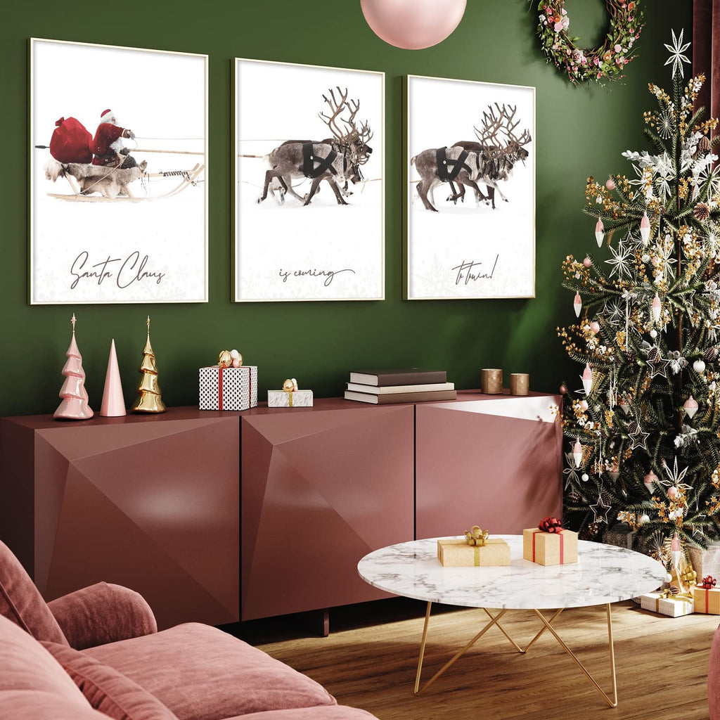 Three framed Christmas art prints depicting Santa Claus in a sleigh and reindeer in the snow, displayed above a console table with Christmas decorations.