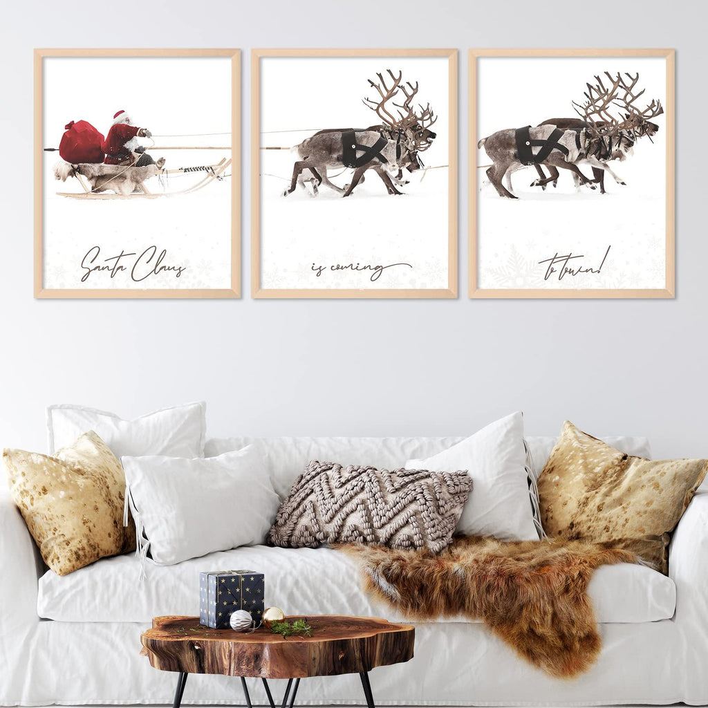Three framed Christmas art prints depicting Santa Claus in a sleigh and reindeer in the snow, displayed above a white sofa.
