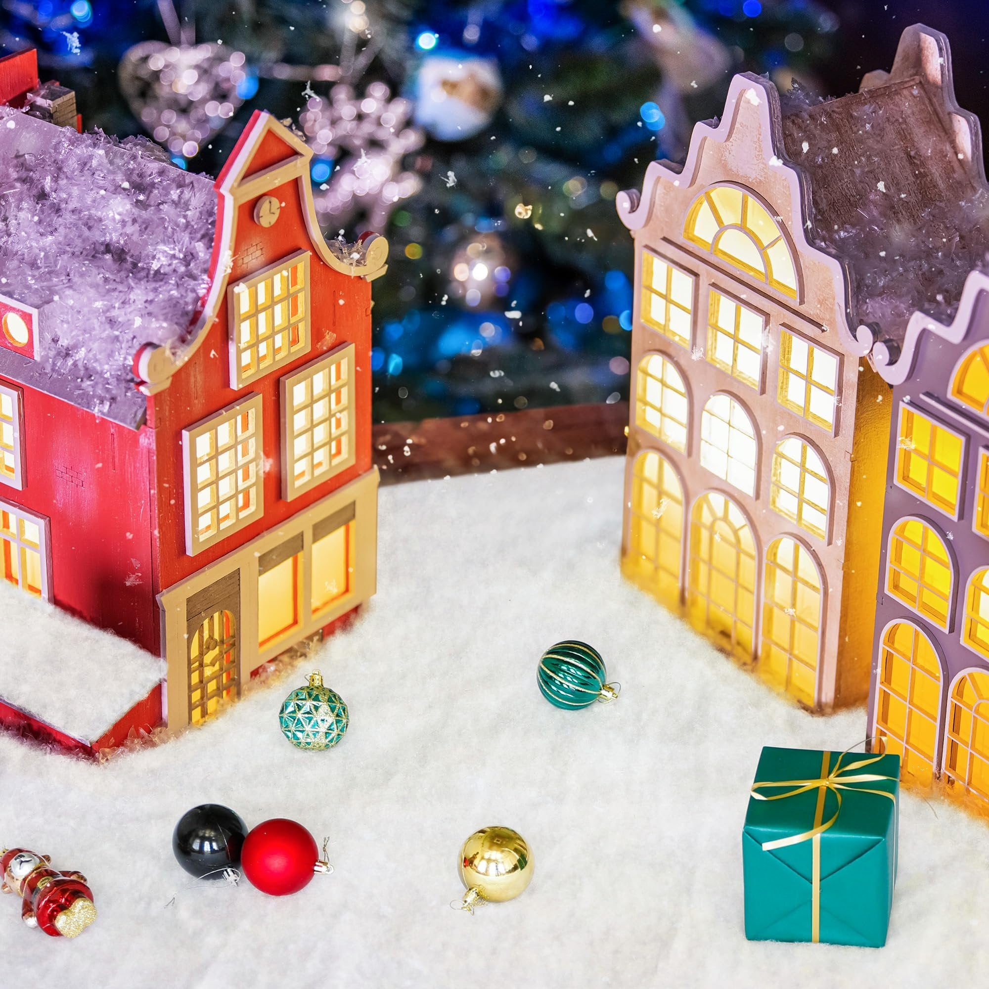 Christmas village scene with illuminated houses, ornaments, and a gift box on artificial snow.
