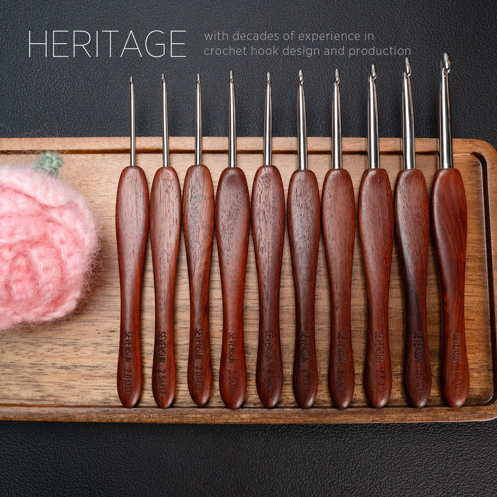 Set of ergonomic crochet hooks with red sandalwood handles and stainless steel tips on a wooden tray next to pink yarn.