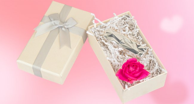 Open gift box with crinkle cut paper filler, a silver lipstick, and a pink rose on a pink background with a faint heart overlay.