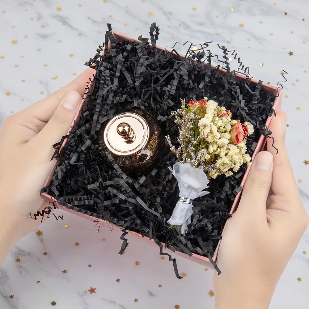 Hands holding a pink gift box filled with black crinkle paper, a gold jar, and a small dried flower bouquet.