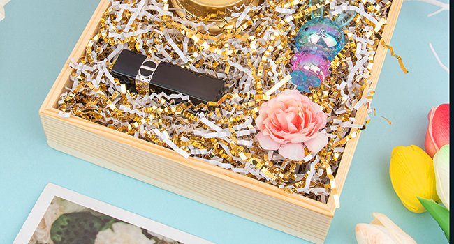 Wooden gift box filled with white and gold crinkle cut paper shred, a black lipstick, a decorative bunny, and a pink rose.