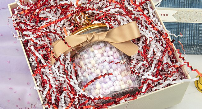 Gift box filled with white and red crinkle cut paper shred, featuring a glass jar with colorful candies tied with a gold ribbon.