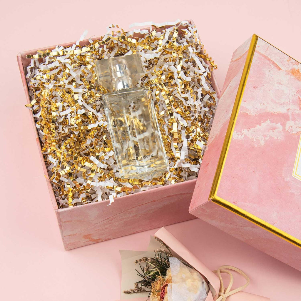 Open gift box filled with white and gold crinkle cut paper shred, with a perfume bottle inside.