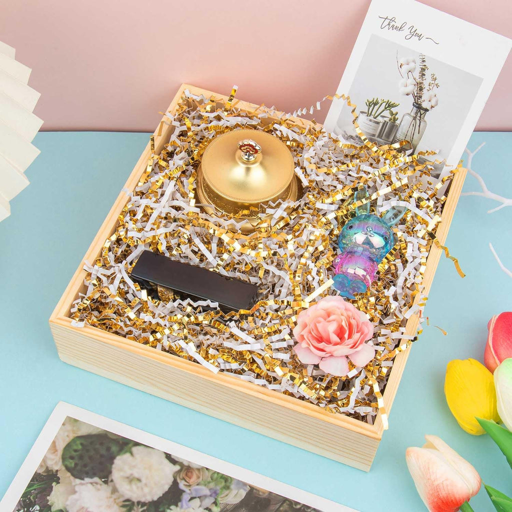 Wooden gift box filled with white and gold crinkle cut paper shred, a gold bell, a black rectangular object, a pink rose, and a colorful bunny charm.