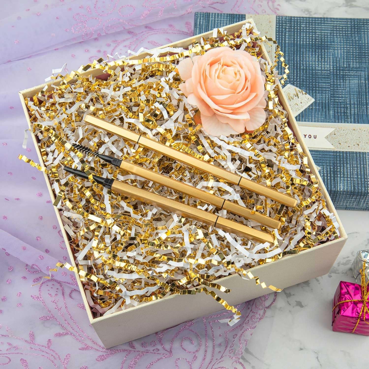 Gift box filled with white and gold crinkle cut paper shred, three gold makeup brushes, and a peach rose.