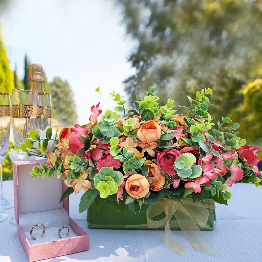 Artificial flower arrangement with eucalyptus and succulents in a green vase, next to a ring box with two rings and champagne glasses.