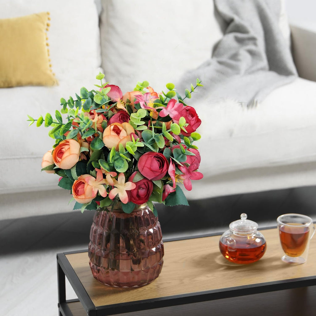 Artificial peony and eucalyptus flower arrangement in a glass vase on a table with tea.