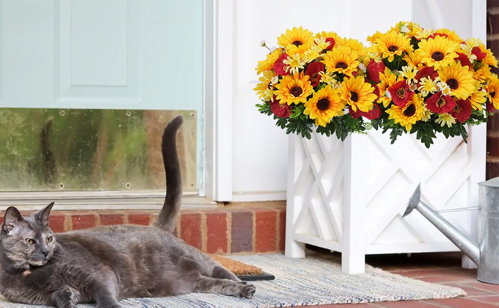 Faux sunflower, daisy, and poppy floral arrangement in a white planter with a cat lying on a rug by a door.