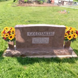 Goldsmith headstone with two vases of faux sunflowers and red flowers on either side, set in a grassy cemetery.