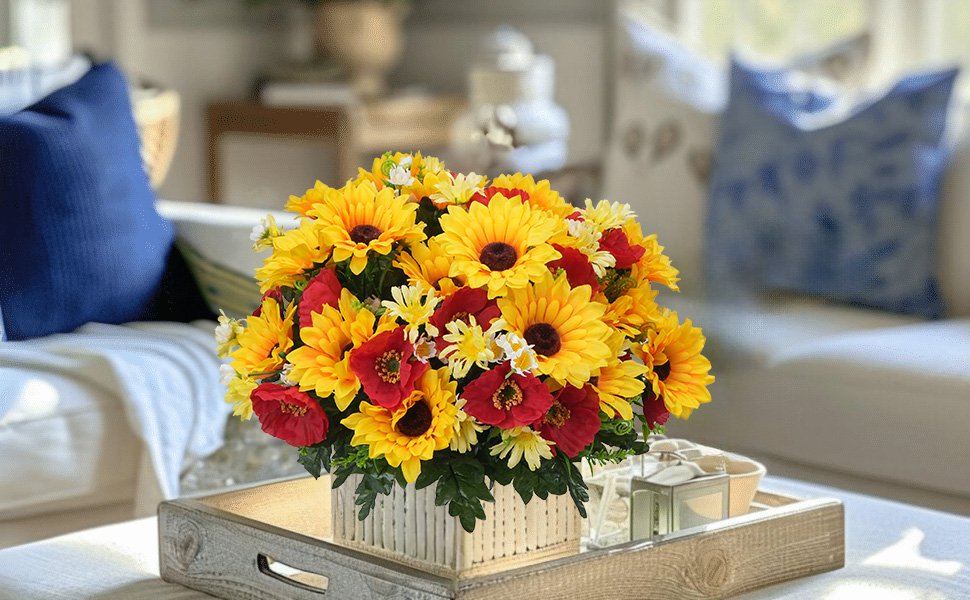 Faux sunflower, daisy, and poppy floral arrangement in a square planter on a wooden tray, set on a table with pillows in the background.