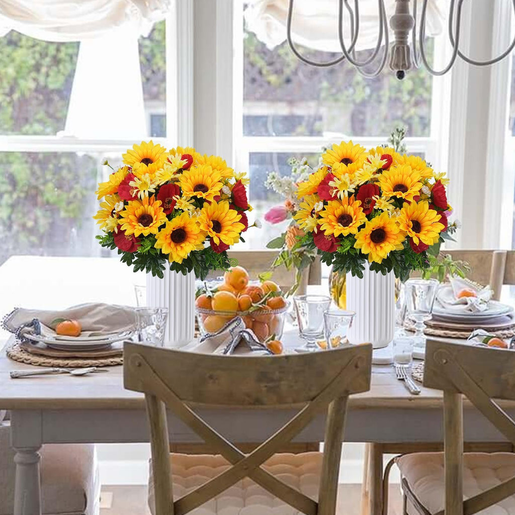 Two bouquets of faux sunflowers, daisies, and poppies in white vases on a dining table set for a meal.