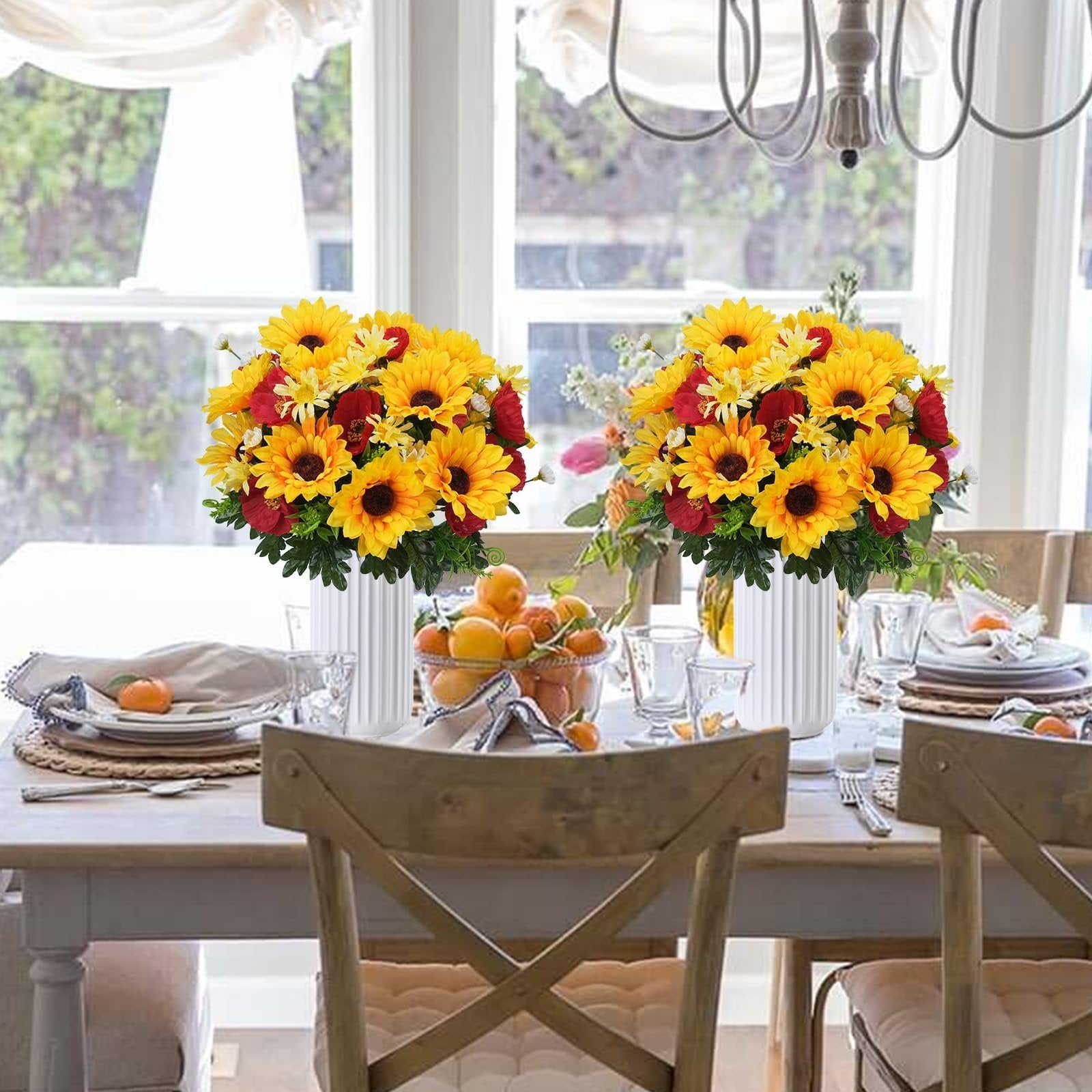 Two bouquets of faux sunflowers, daisies, and poppies in white vases on a dining table set for a meal.