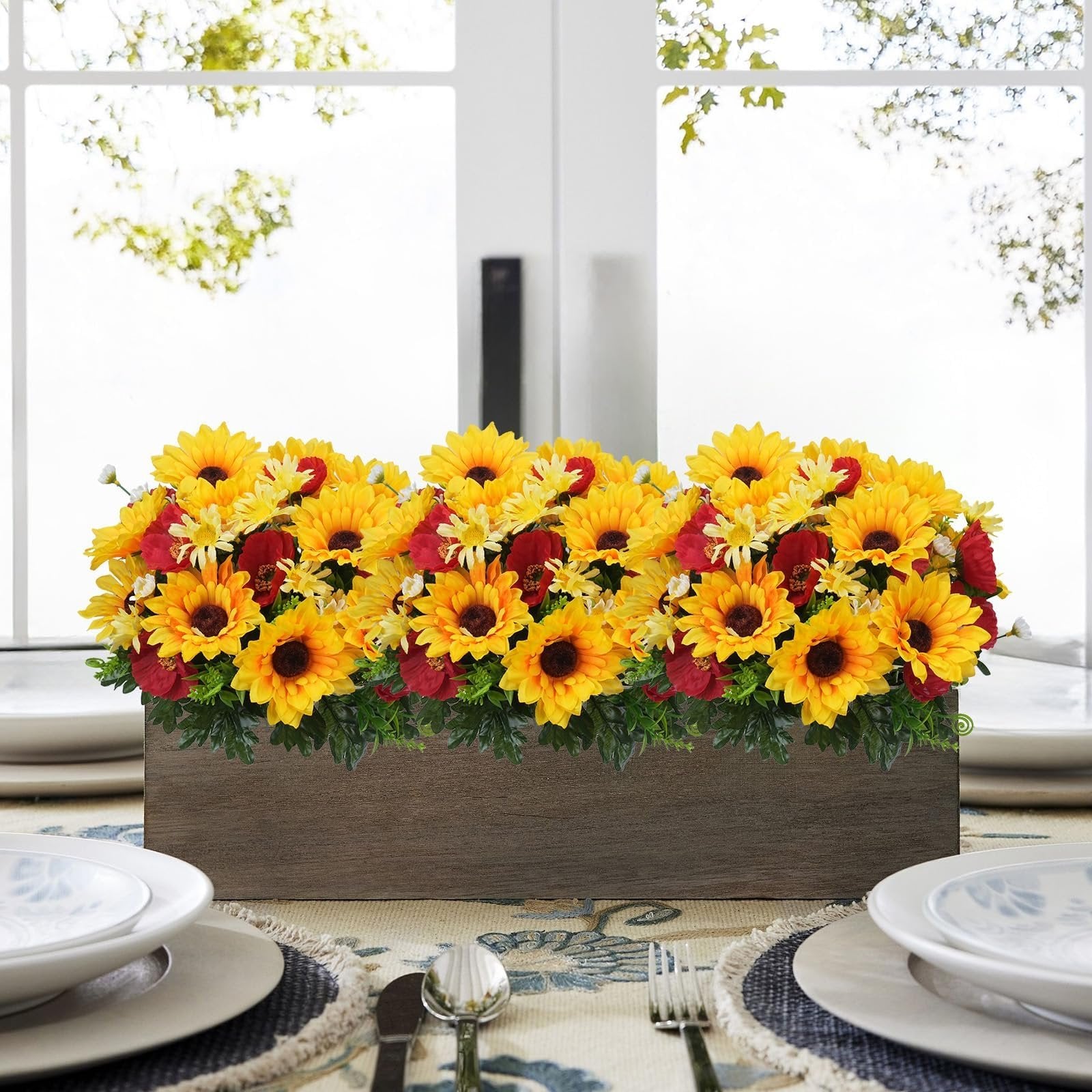 Faux sunflower, daisy, and poppy floral arrangement in a wooden box on a dining table setting.